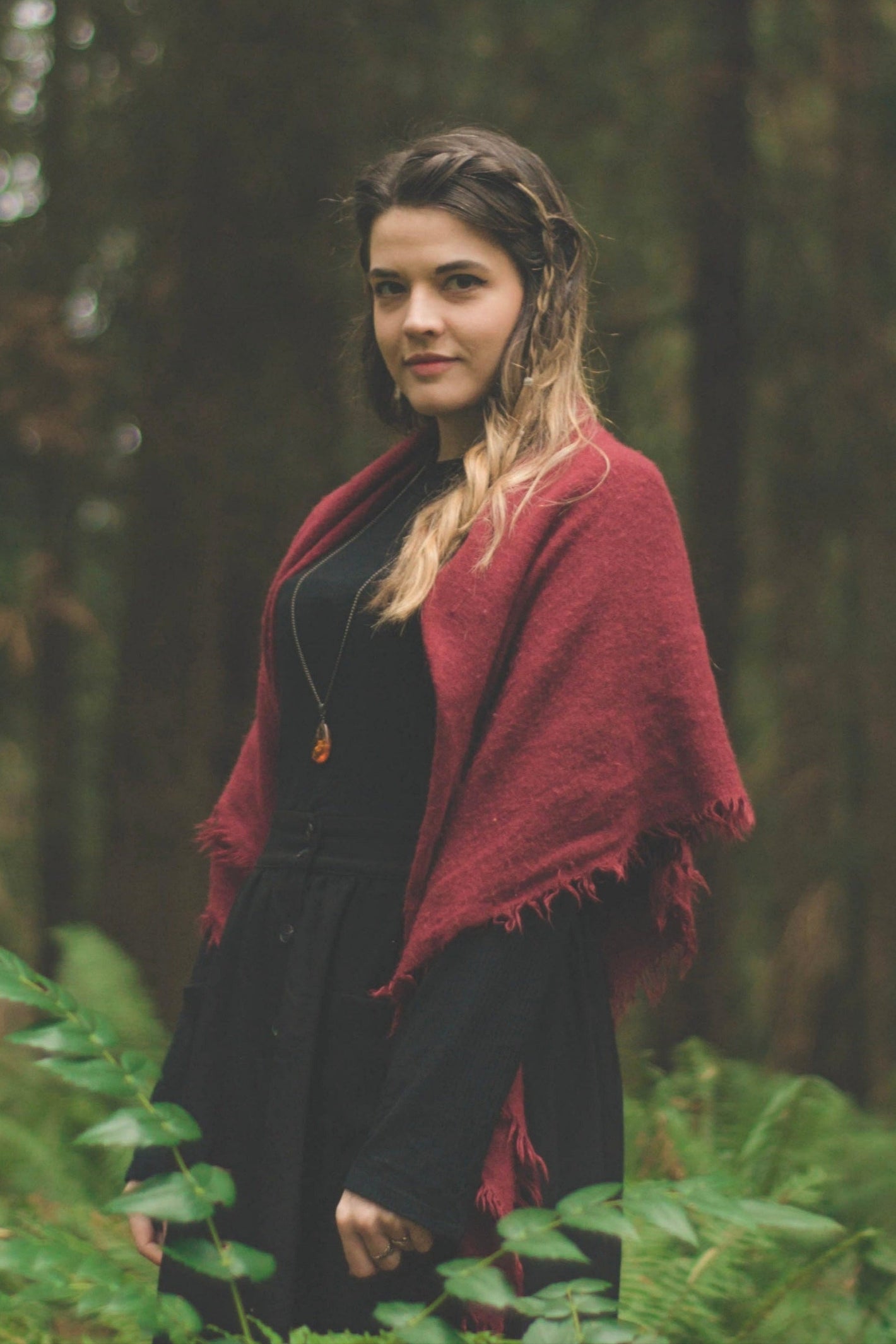 A long-haired forest witch in a black dress and red shawl, standing amongst redwood trees, ferns and oregon grape vines